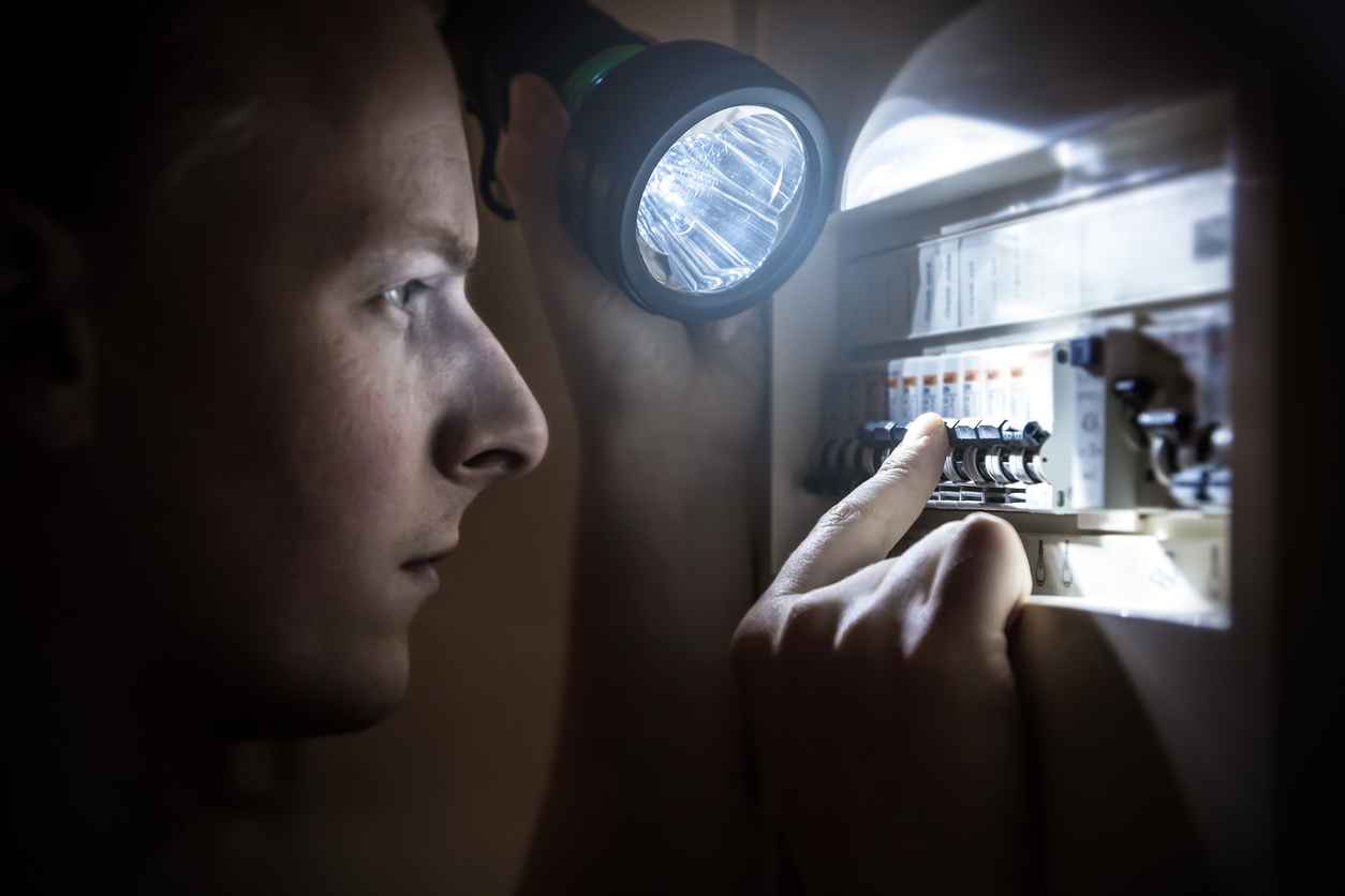 Horizontal composition photography of a young man, specialized technician or just home man, trying to restore power after electricity outage in the dark with his light. He is looking on the circuit breaker, and try to find the right button to operate.